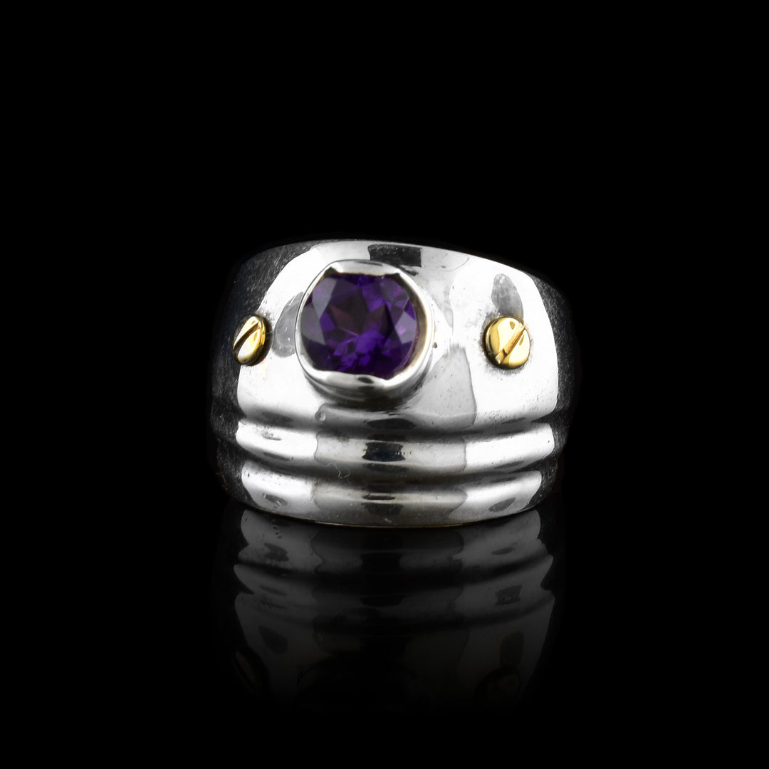 This sterling silver amethyst solitaire ring has a wide band and is tapered. At the top of the ring is an embedded amethyst gem with a 14K gold stud on both sides.