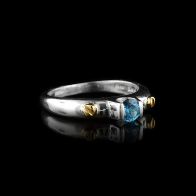 This blue topaz solitaire ring has a thin band made from sterling silver with a blue topaz gem set at the top of it. The gem is displayed above the band with two silver pieces at its side holding it up. There are 14K gold studs on the band to either side of the gem.