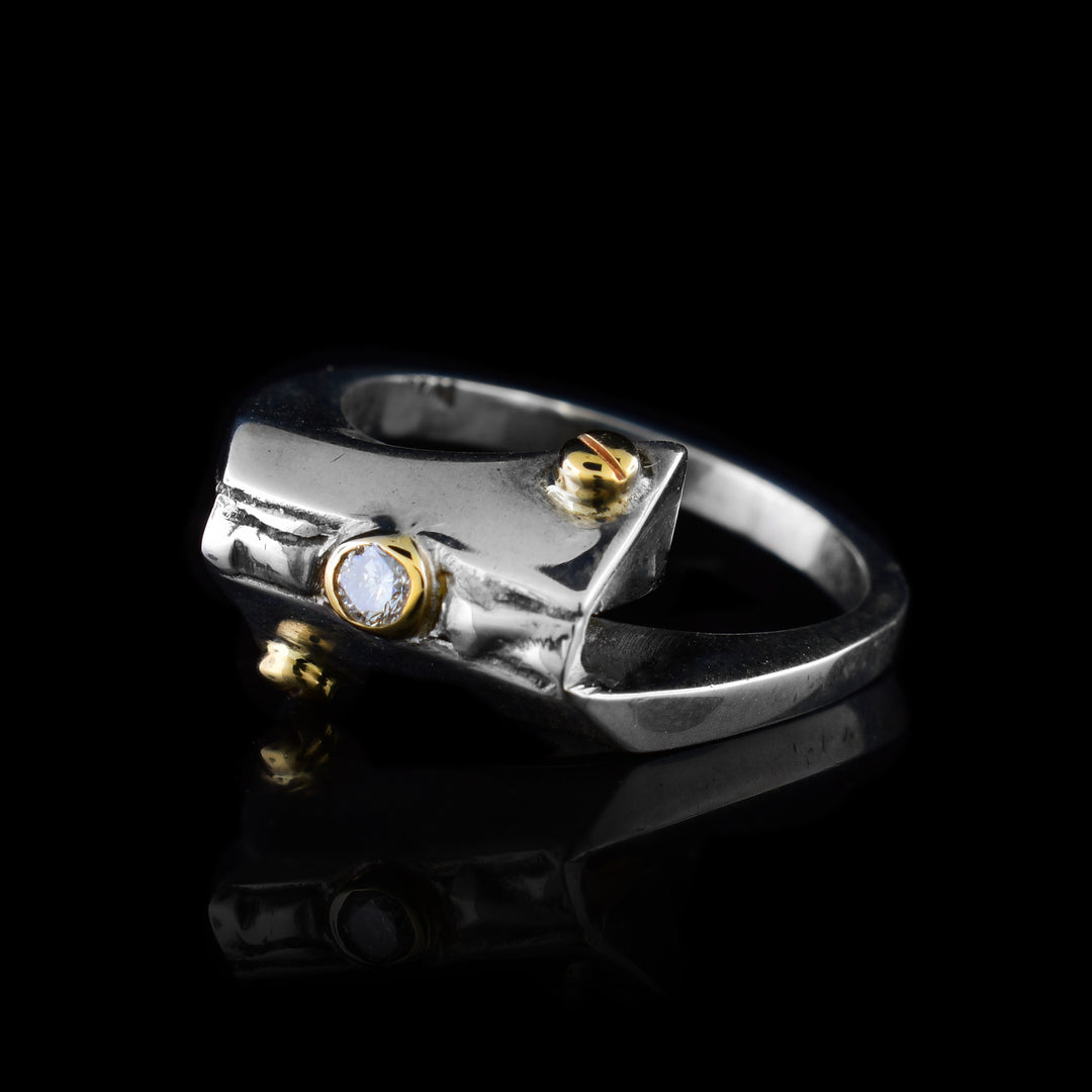 This sterling silver diamond ring has a thin band with a rectangular adornment on it. Attached to the adornment are two 14K gold studs and a diamond wrapped in gold.
