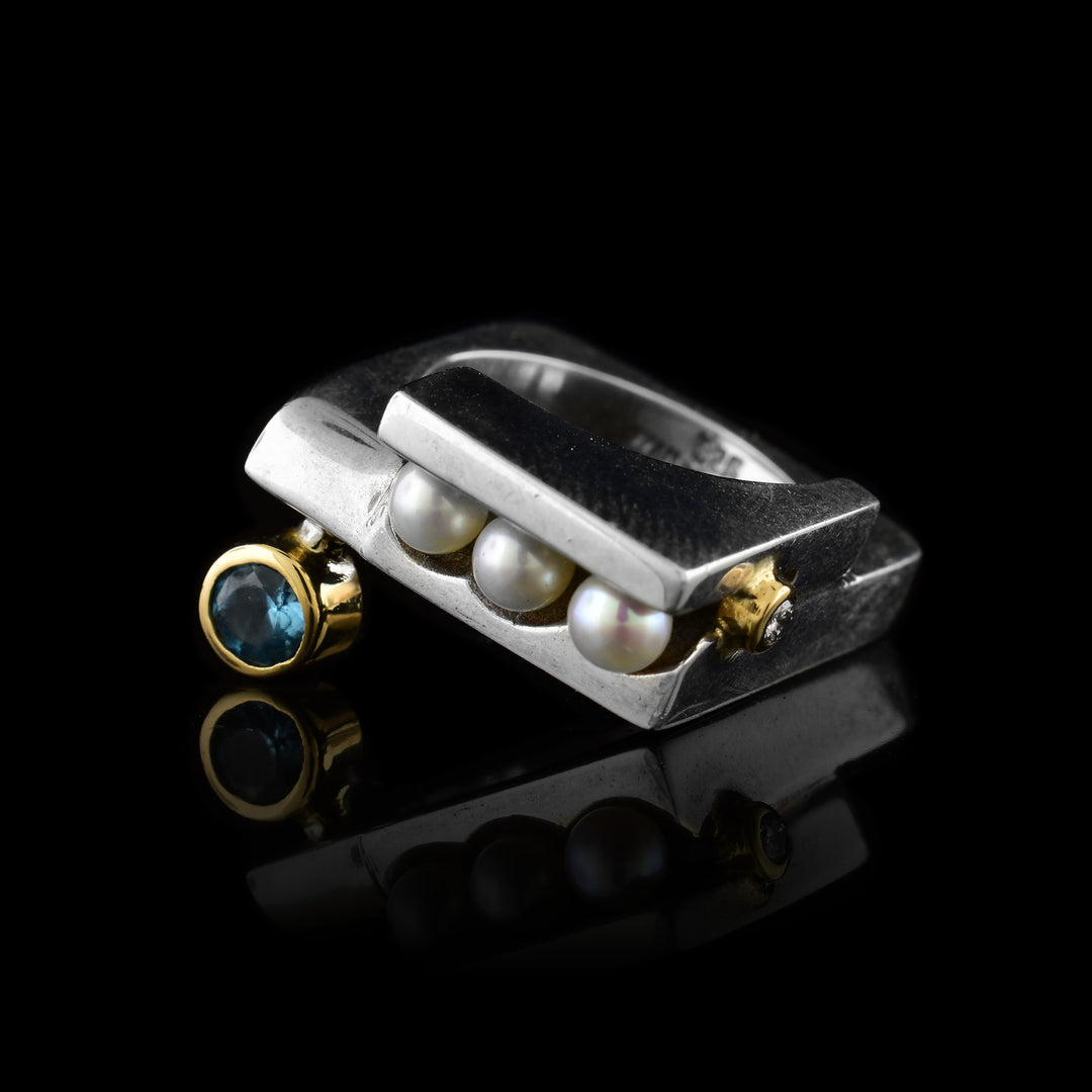 This sterling silver ring has a square shape. Attached into the signet are three freshwater pearls. Attached to the ring with 14K gold is a blue topaz gemstone and a diamond on either end of the ring.