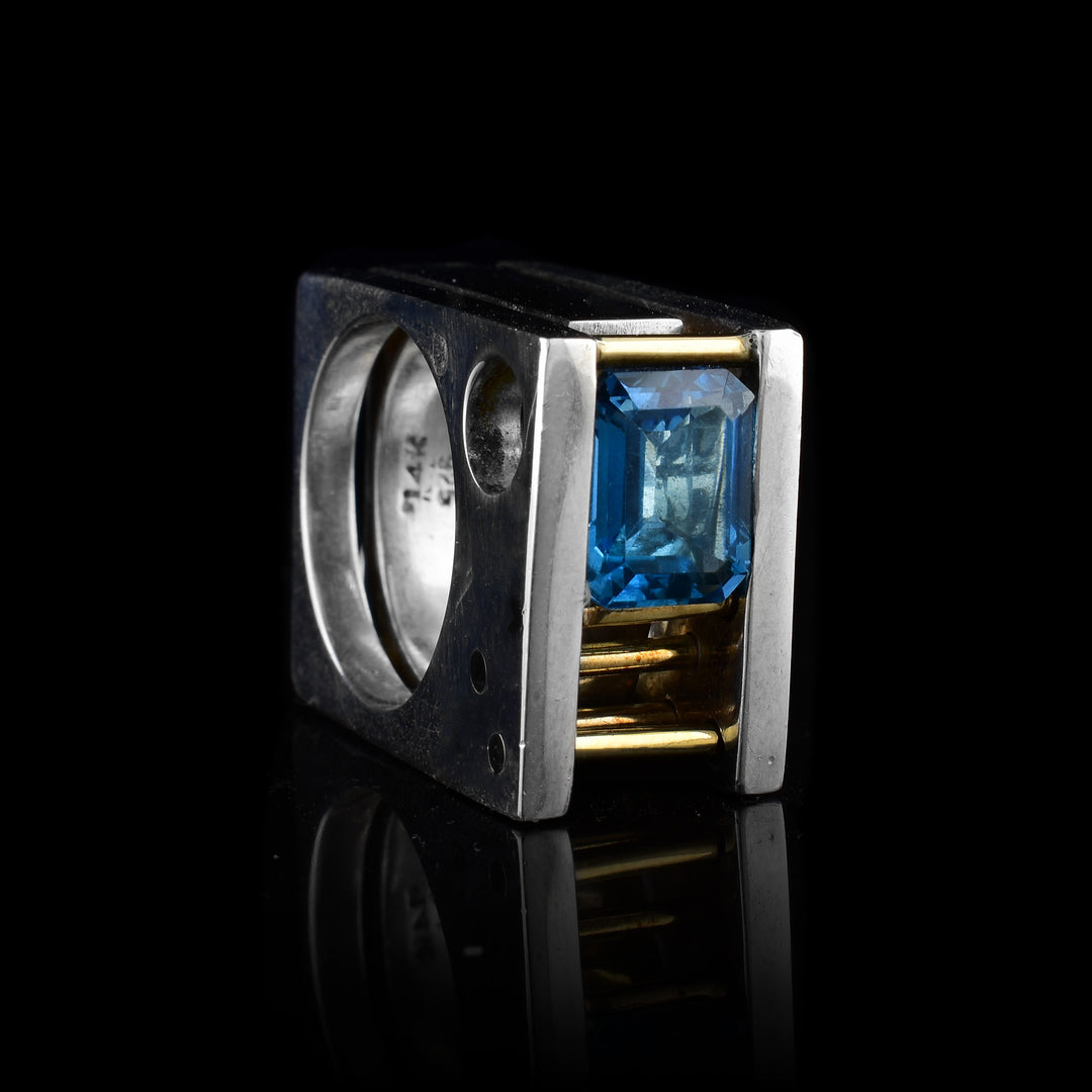 This abstract, square ring has three sterling silver layers with a blue topaz gemstone between them. By the gemstone, there are five 14K gold rods running through two bands. The entire ring is square shaped.