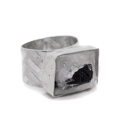 This quartz ring has a bulging rectangular silver casing with clear quartz inside. There is a black tourmaline peeking out of the quartz. The band is wide and has a pattern on it on the outside.