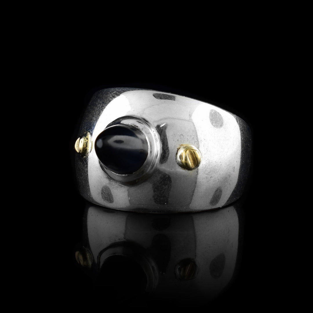 This wide band solitaire ring is made from sterling silver and has a side band that tapers slightly in the back. There is a large, cone shaped black onyx gem in the center that points outwards from the band. There are 14K gold studs to either side of it.