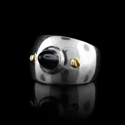 This wide band solitaire ring is made from sterling silver and has a side band that tapers slightly in the back. There is a large, cone shaped black onyx gem in the center that points outwards from the band. There are 14K gold studs to either side of it.