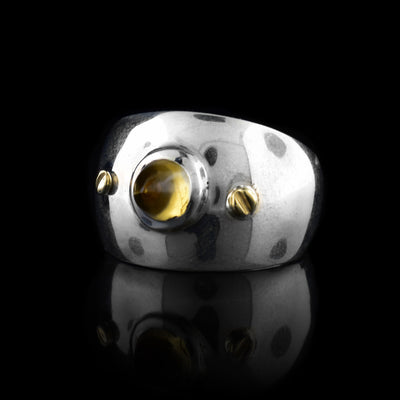 These citrine rings have wide, tapered and sterling silver bands with a large circle shaped citrine gem embossed in the center of the solitaire ring. There are two 14K gold studs placed on either side of the gem.