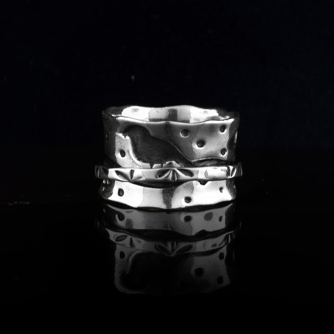 This sterling silver spinner ring has a wide band with polka dots of oxidized silver across the surface of it. There is a very thin band wrapping around the middle of the band with small, leaf designs on it. The center band can be spun around the piece.