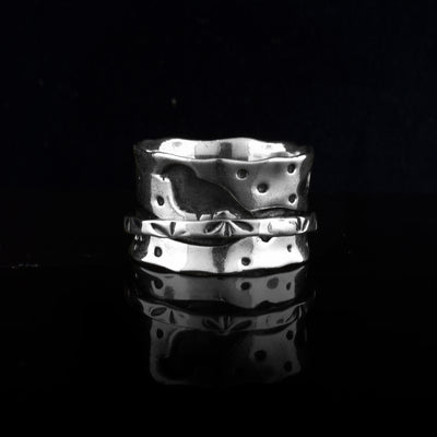 This sterling silver spinner ring has a wide band with polka dots of oxidized silver across the surface of it. There is a very thin band wrapping around the middle of the band with small, leaf designs on it. The center band can be spun around the piece.