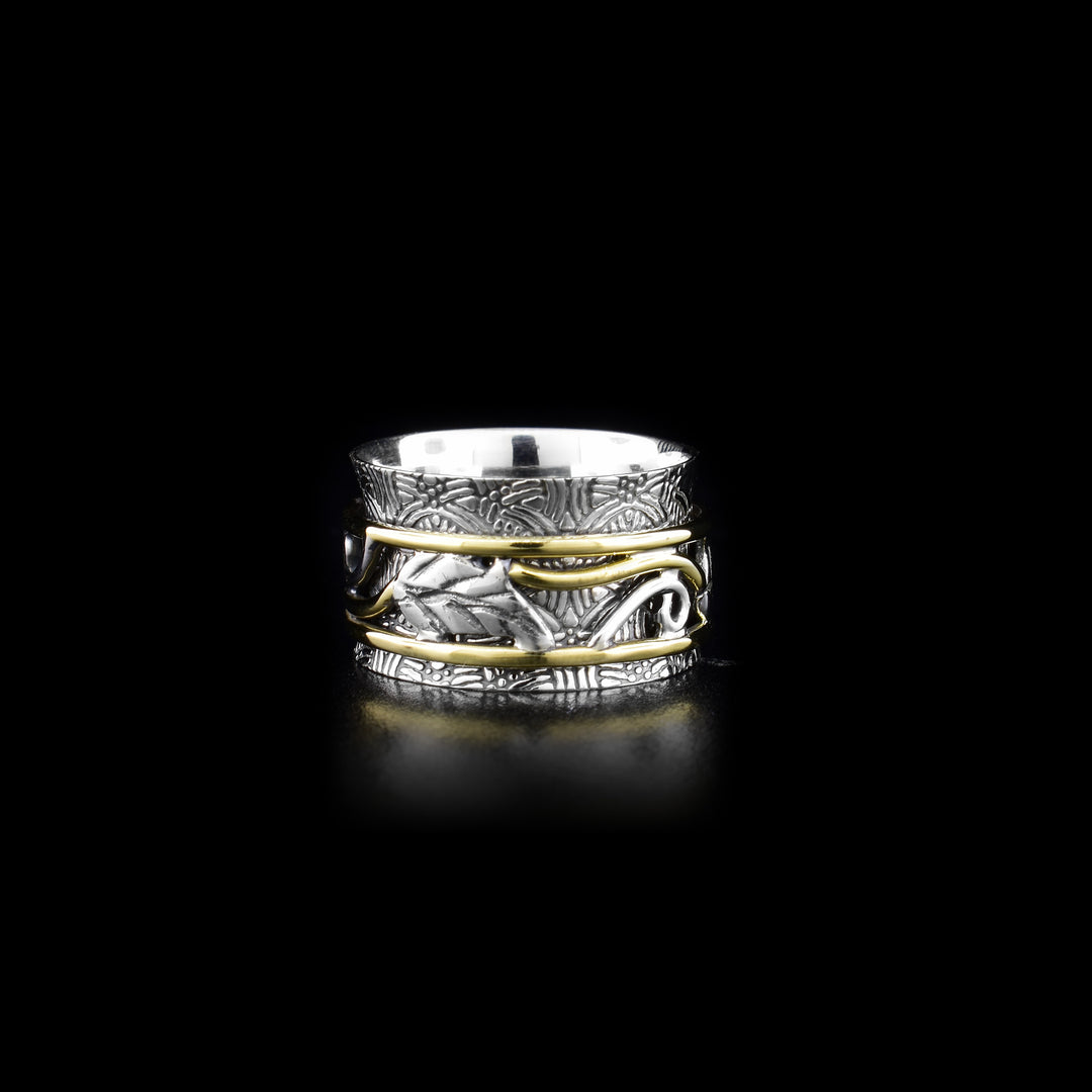 This spinner ring has a wide, sterling silver band with textured designs made across the surface of it. The center part of the ring is framed by brass wires and has leaf designs on it and decorative brass wires. The center band can be spun around the piece.