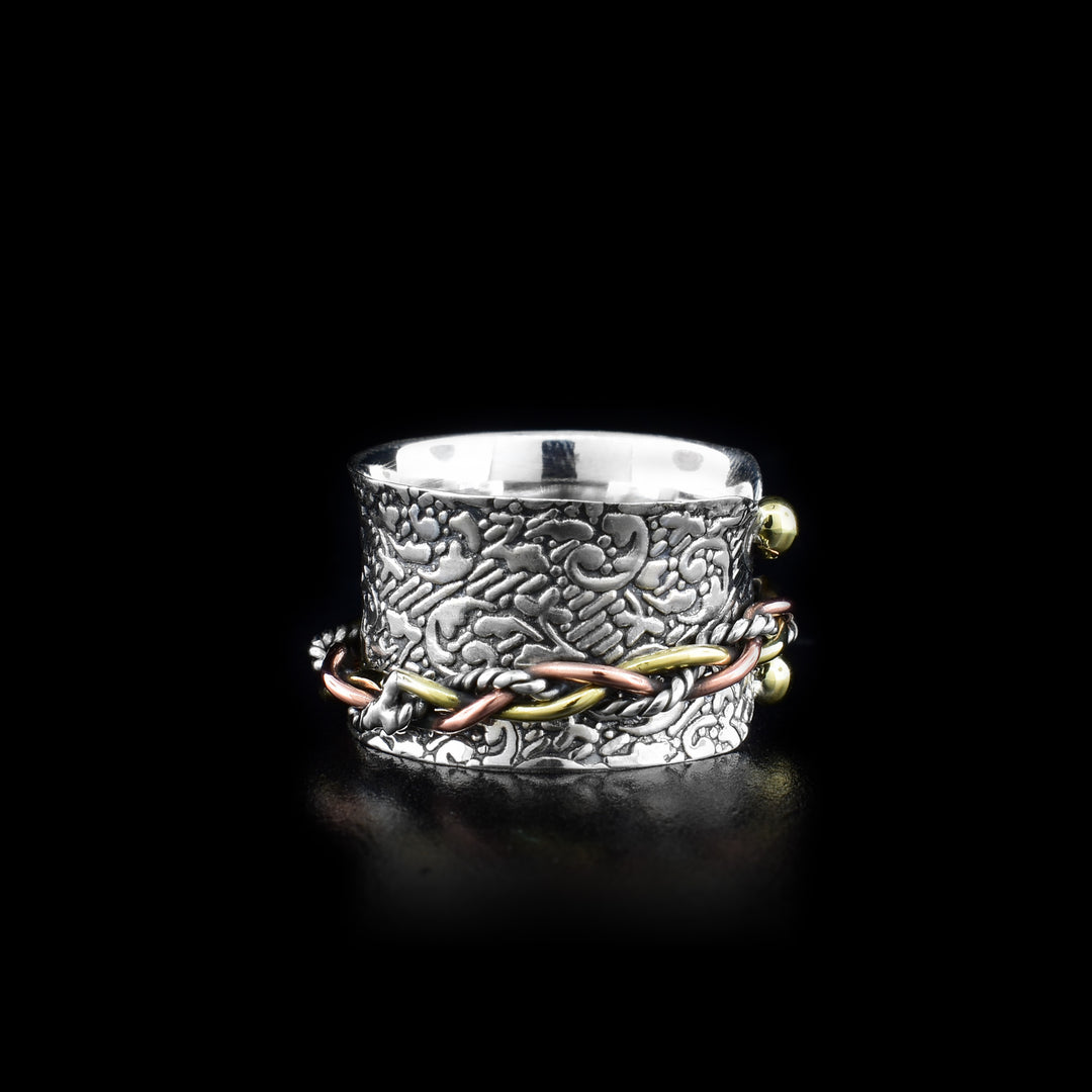 This spinning chain ring has a wide, sterling silver band that has textured designs made on the surface of it.  There is a braided chain made from silver and copper that wraps around the band and can be spun around the piece. There is a small gap in the front of the band with two brass beads connected to band. The outside of the band flaires out slightly.