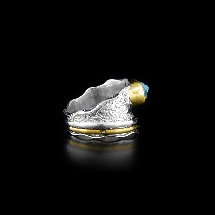 This spinner ring is made from sterling silver, brass and apatite gemstone. There is a wavy, tapered and silver band with designs imprinted on it and the large gem placed on top of the band. There is a silver hoop and brass hoop on the band that can be spun around the piece.