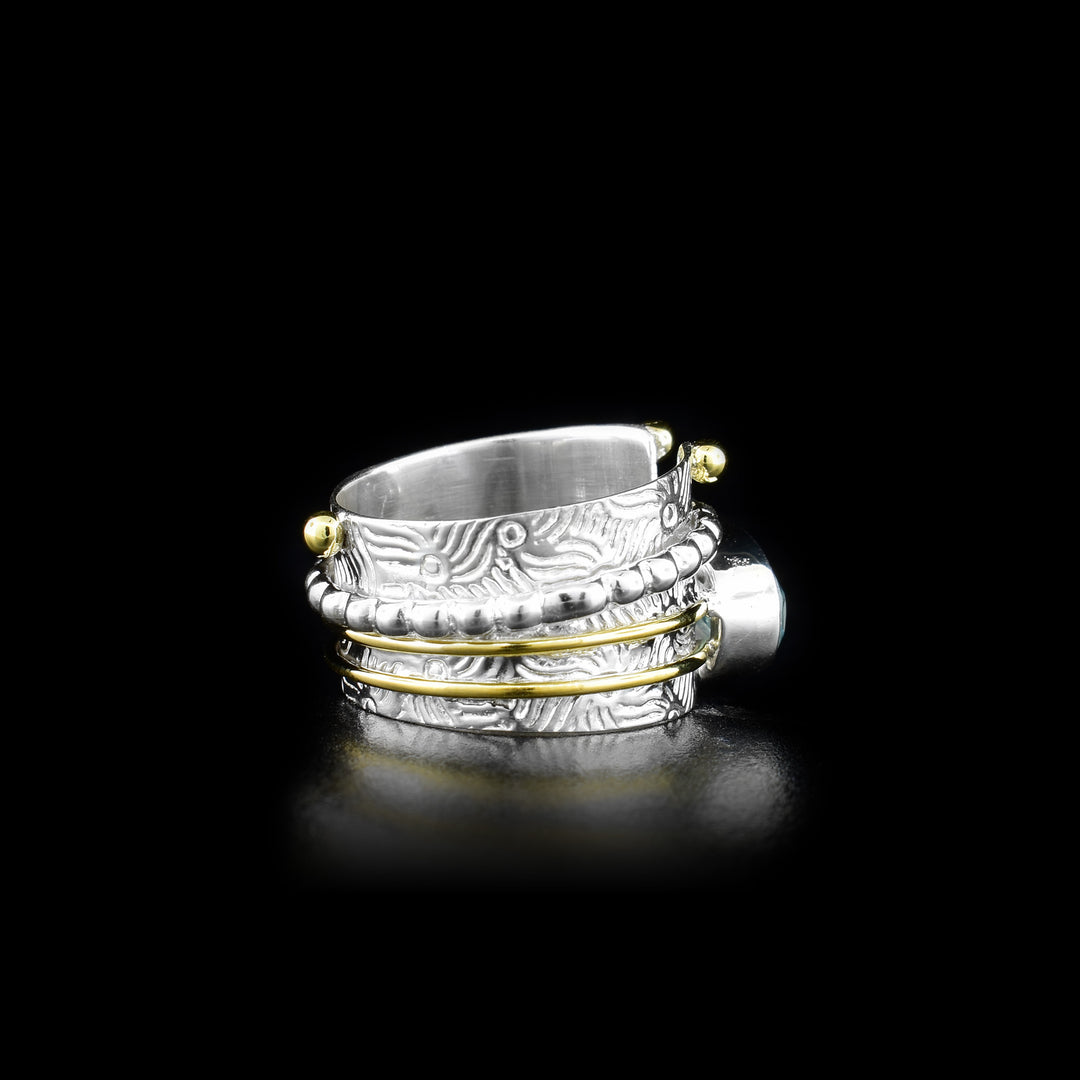 This stunning spinner ring has a sterling silver band that has designs made on the surface of it, a split on the top of it and it tapers slightly. There is a large, ovoid blue topaz gem that is held obove the band and has two brass wires wrapping around the band extending from the gem. There is a silver ring wrapped around the band that has circular textures and can be spun around the piece.