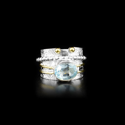 This stunning spinner ring has a sterling silver band that has designs made on the surface of it, a split on the top of it and it tapers slightly. There is a large, ovoid blue topaz gem that is held obove the band and has two brass wires wrapping around the band extending from the gem. There is a silver ring wrapped around the band that has circular textures and can be spun around the piece.