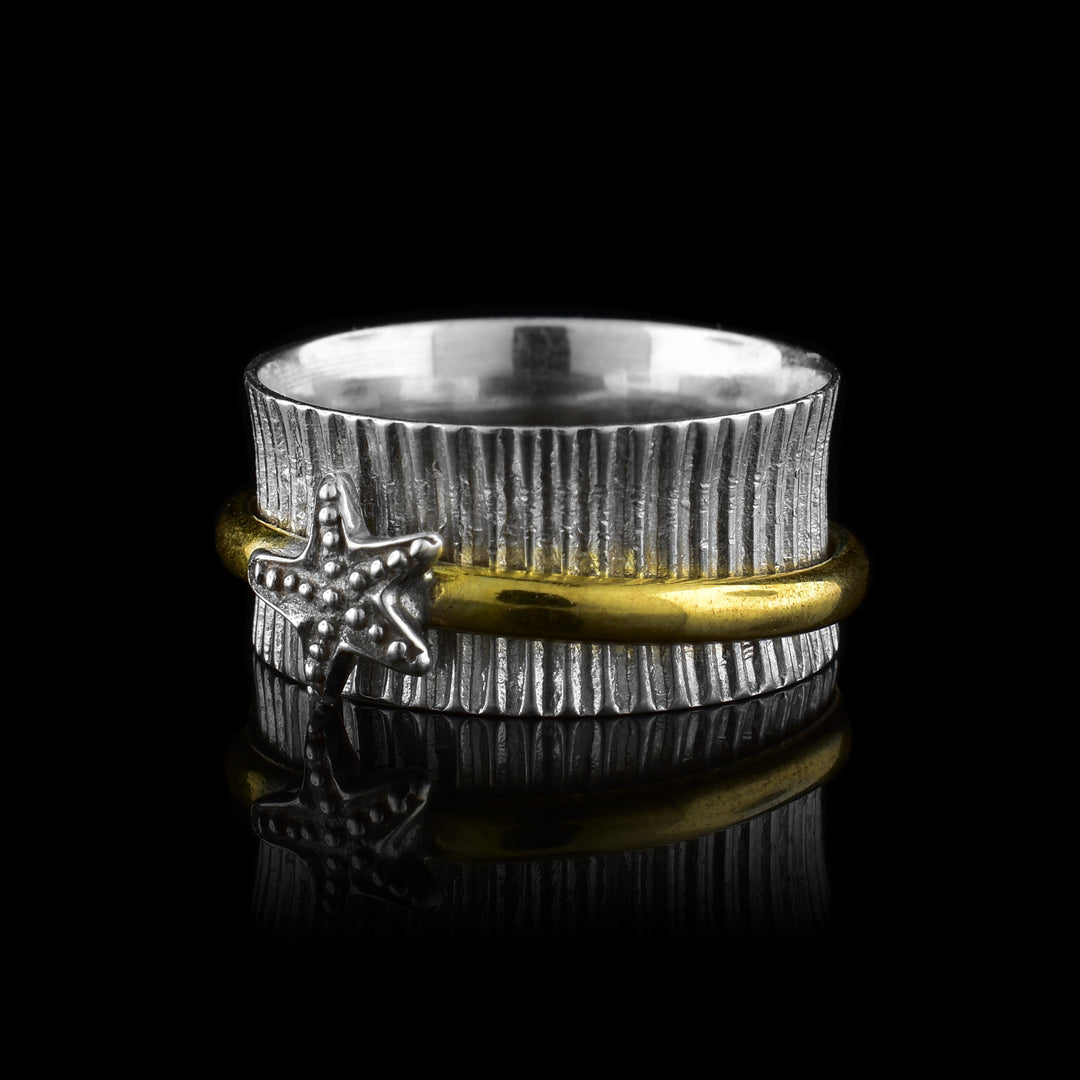 This ladies spinner ring has a wide band made from sterling silver and has vertical ridges made around the entire band. There is a thin, brass band in the enter of the piece that spins around the band. here is a silver starfish on the brass band.