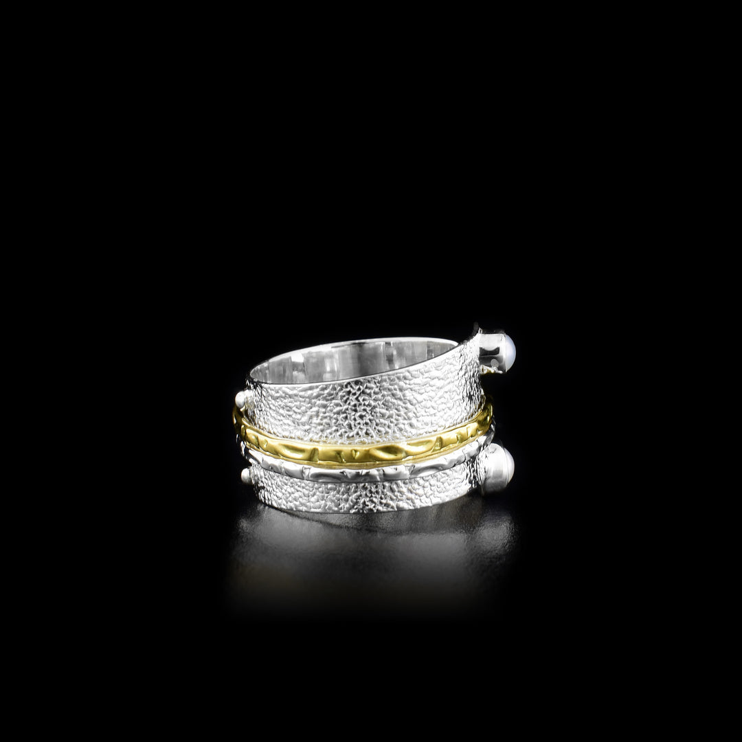 This stunning spinner ring has a wide sterling silver band with a textured surface and two small, round fresh water pearls embossed onto either end of the bands width. There are ribbed textures on a silver and a brass ring that is wrapped around the band. These textures rings can be spun around the band.