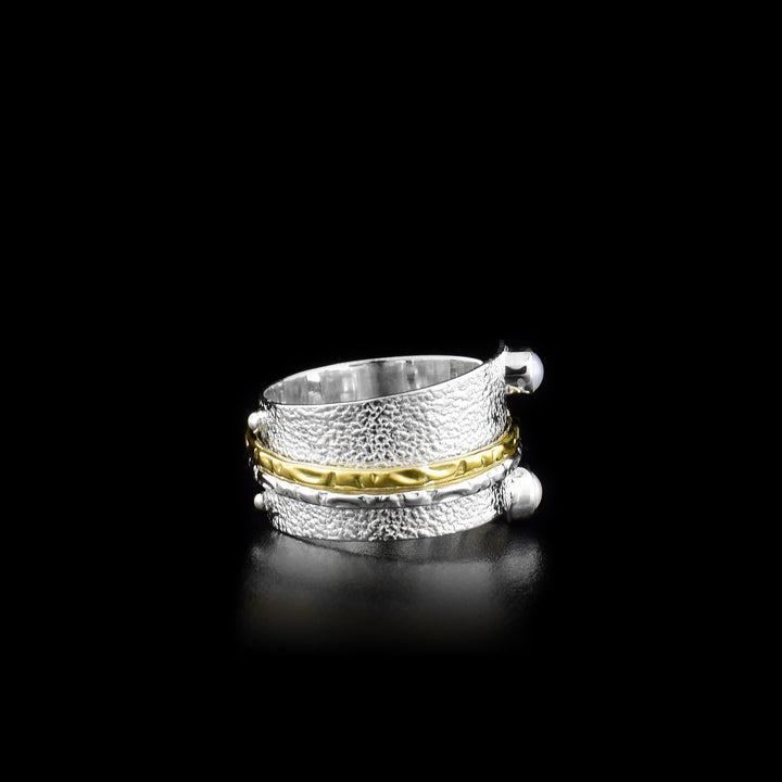 This stunning spinner ring has a wide sterling silver band with a textured surface and two small, round fresh water pearls embossed onto either end of the bands width. There are ribbed textures on a silver and a brass ring that is wrapped around the band. These textures rings can be spun around the band.