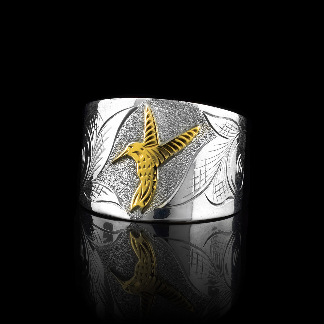 This sterling silver and gold Hummingbird ring has a wide band made from sterling silver that tapers in the back. The Hummingbird is done in 10K gold and has its wings out stretched in the center of the piece. There are leaf designs around the bird.