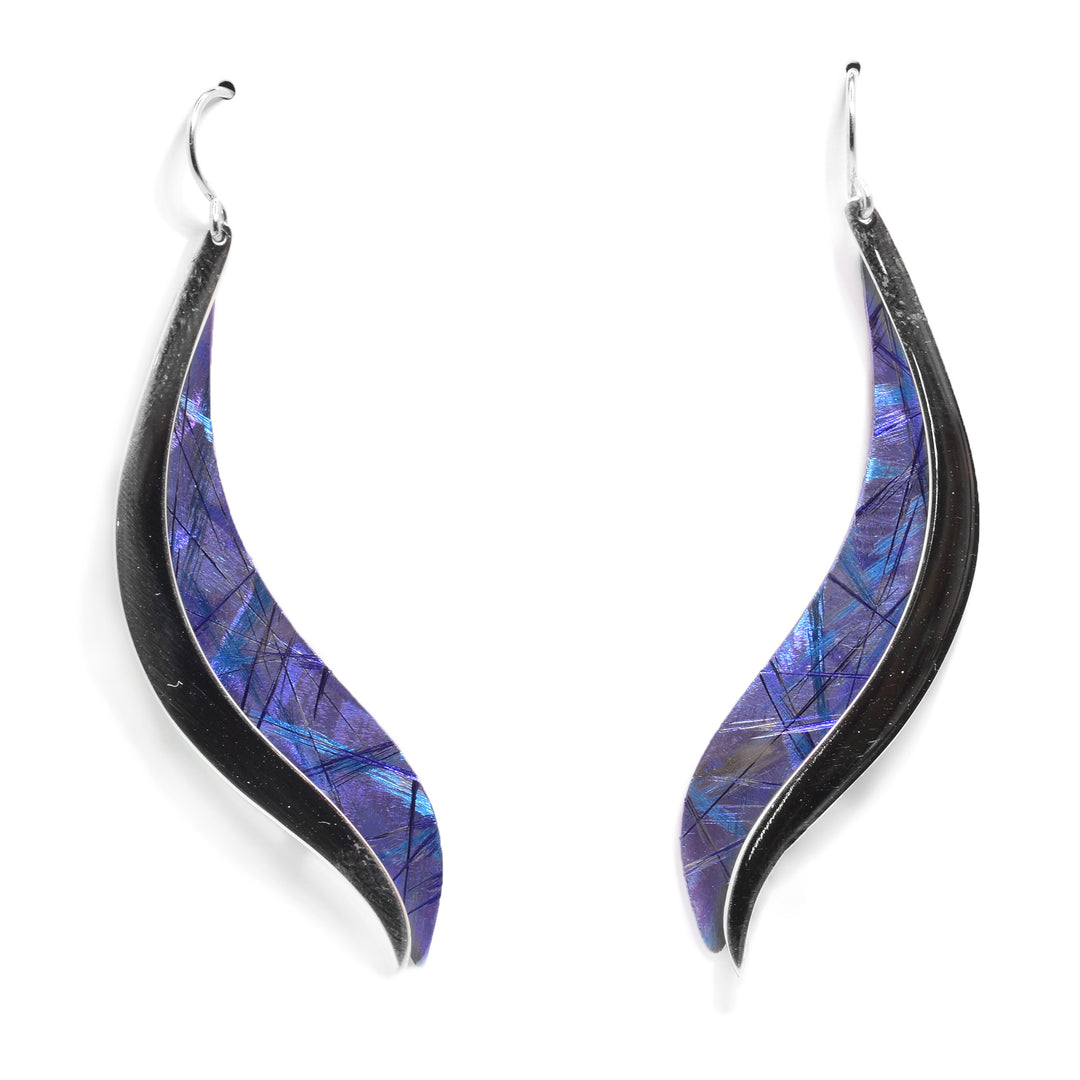 These titanium drop earrings have long wavy hangs made from titanium. There are purple and blue designs on the hangs achieved by anodizing the material. There is a sterling silver band going across one end of the hangs.