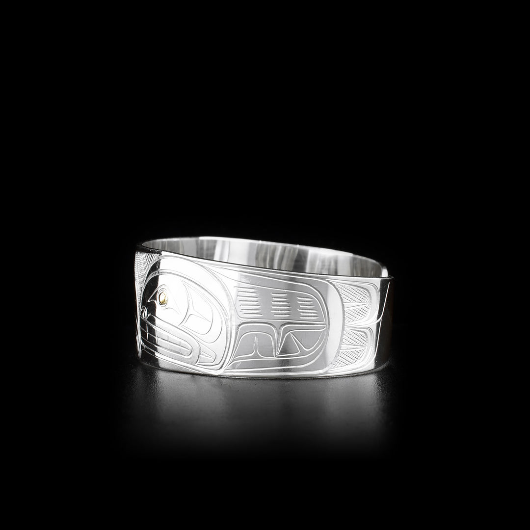 This silver and gold bracelet is made from sterling silver and 14K gold and has a wide band with a gap in the back. There are depictions of the Raven and the Orca carved into the surface of the band. The eyes of the legends are made from 14K gold.