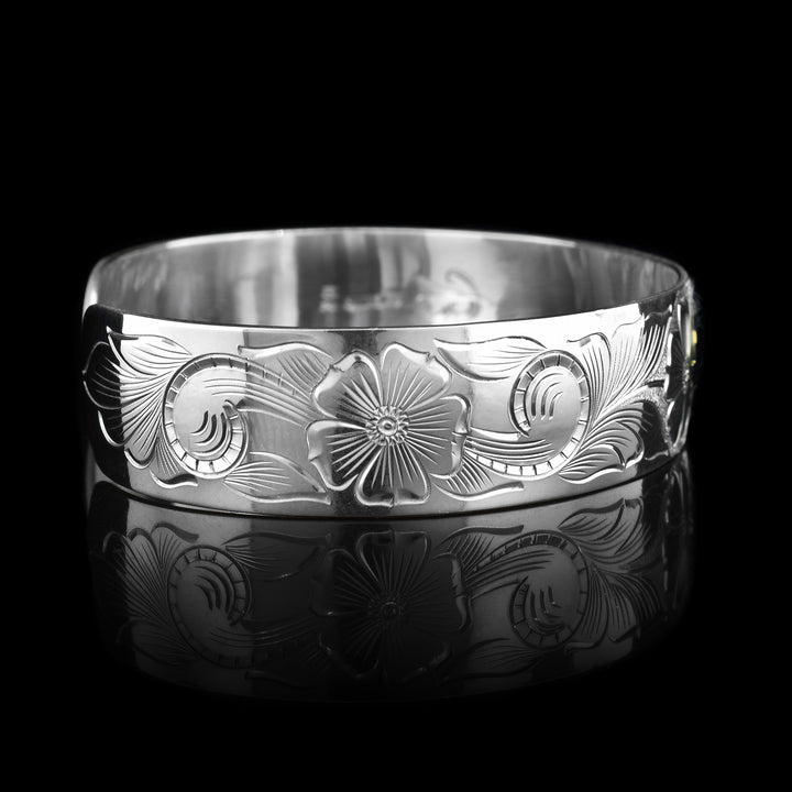 This sterling silver hummingbird bracelet cuff has a wide band with carvings that depict flowers and a hummingbird. The hummingbird is made of 10K gold.