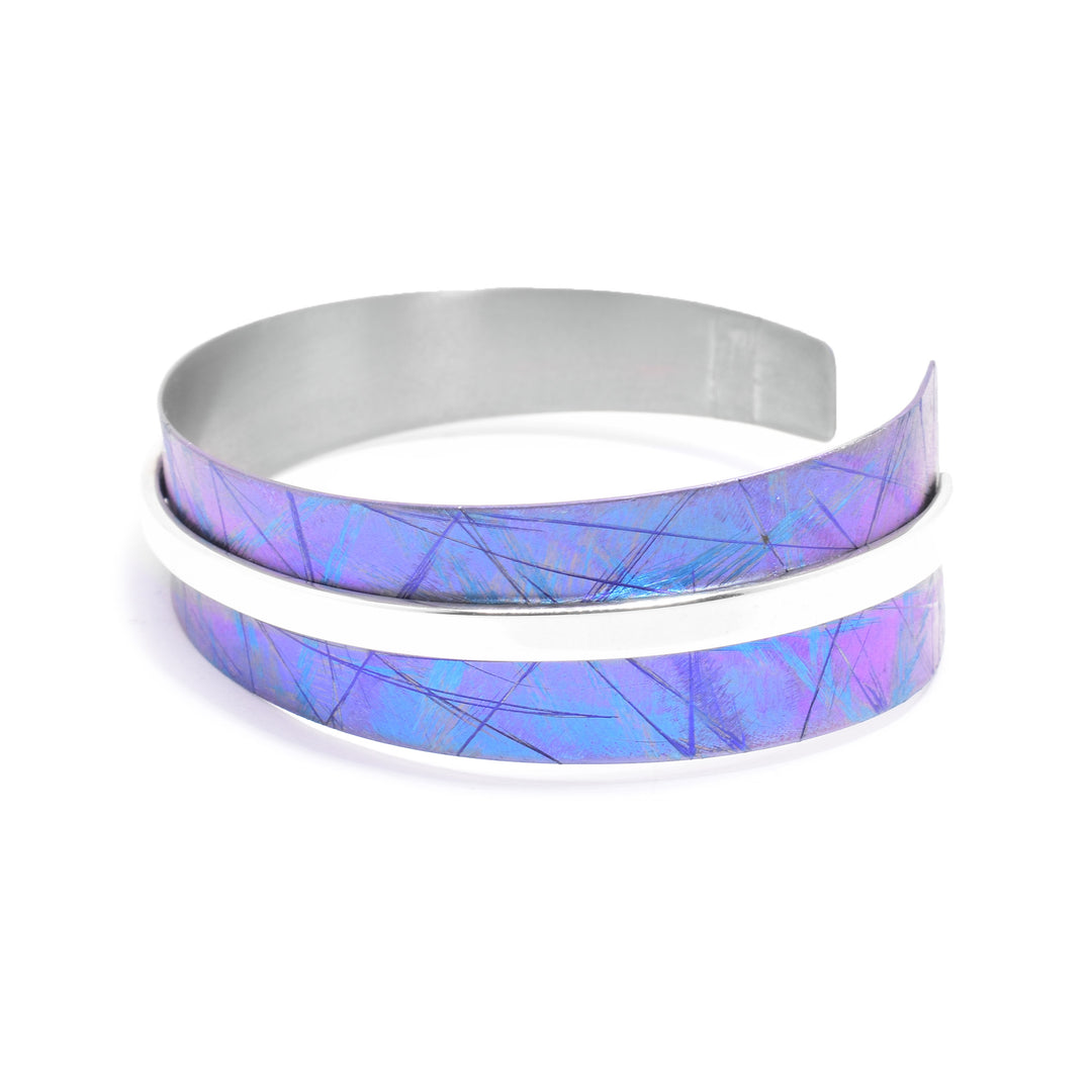 This anodized titanium bracelet has a single, tapered band with a gap in the back and a sterling silver strip running through the middle of the band. The titanium band features a blue color with colorful designs that have been achieved by anodizing the meatal.