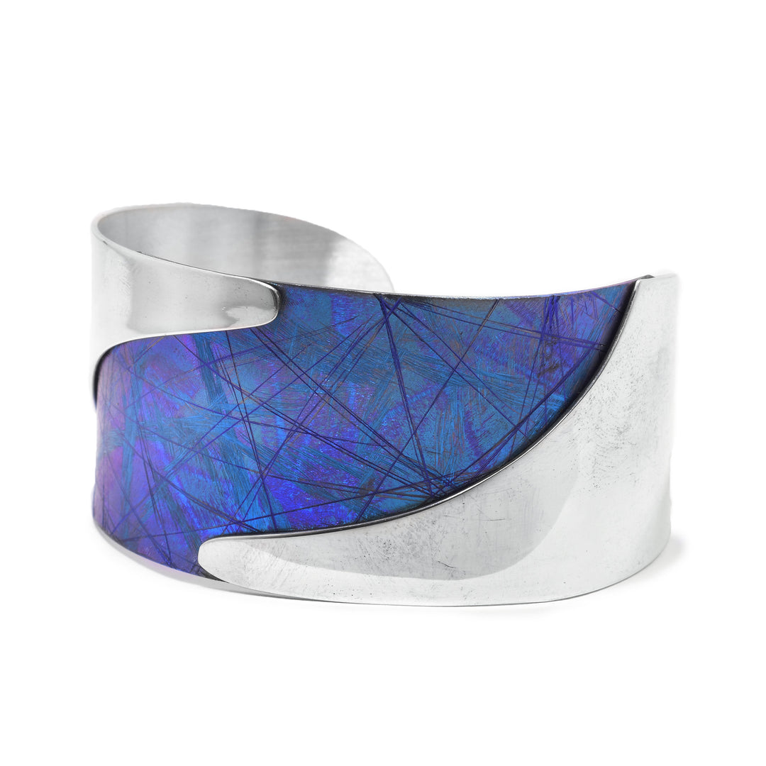 This titanium bracelet is made from sterling silver and anodized titanium and has a single wide band. The titanium is anodized to be blue and has colorful designs, there are wavy, sterling silver highlights running along the bracelet.