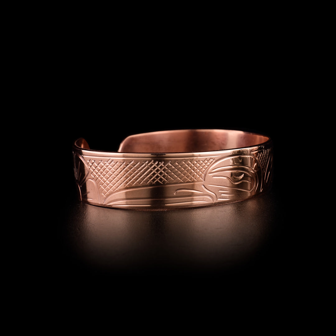 This Hummingbird cuff bracelet has a single band with a gap in the back and is made from copper. There is a depiction of the head of the Hummingbird carved at a side profile into the band. The beak is facing a flower on one end and on the other its wing is seen out stretched. There are cross hatching designs in the background of the scene.