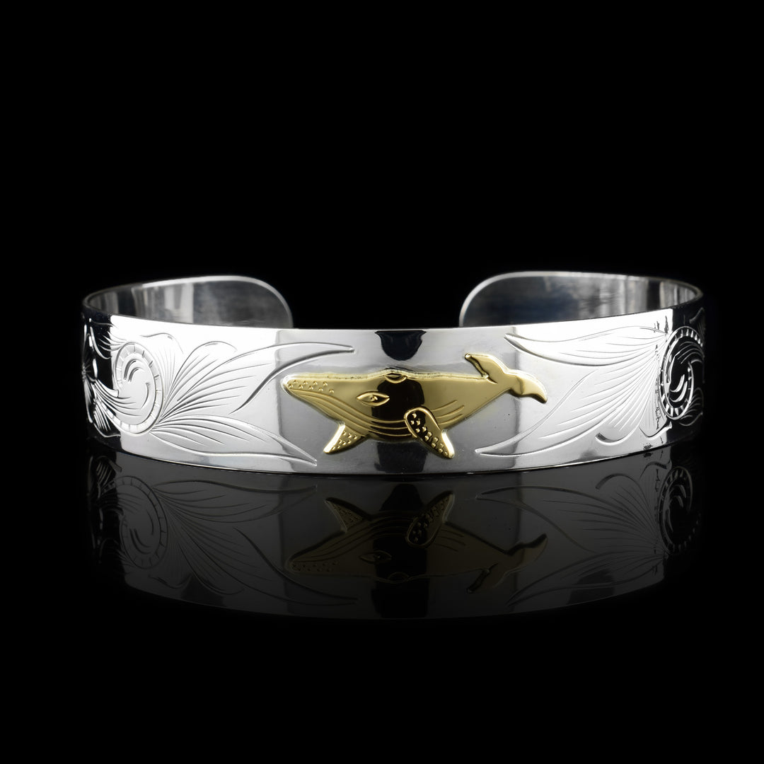 This whale cuff bracelet has a single, sterling silver band with a gap in the back. There is a 10K gold whale in the center of the band with swirling designs carved around it across the band.