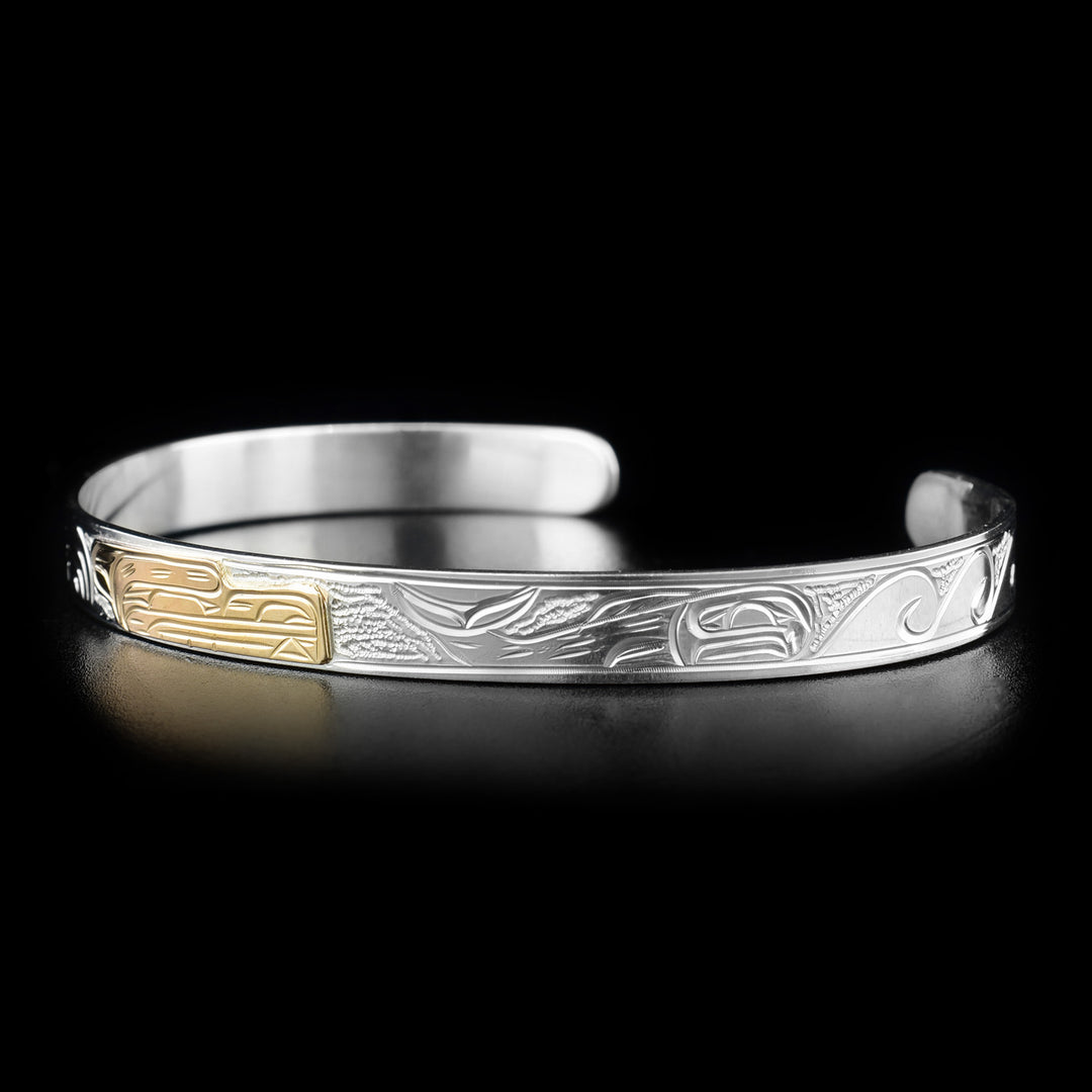 This bear bracelet depicts the head of a bear facing the right in the center of the bracelet made from gold. To its right is a full-bodied salmon with waves in front of it. Background is stippled.