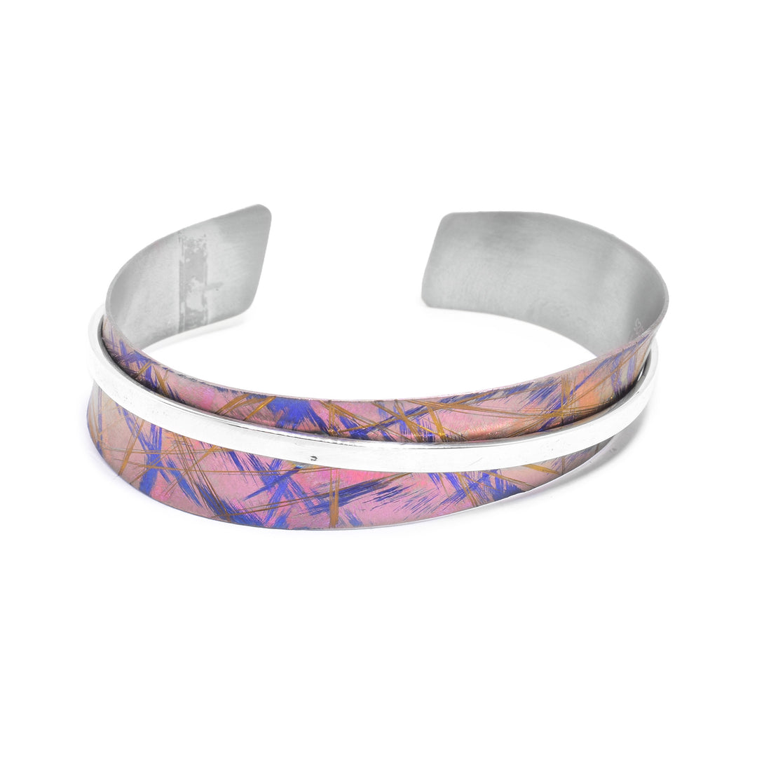 This titanium bangle bracelet has a titanium band with a gap in the back. There are pink, blue and gold designs on the titanium. There is a thin, sterling silver band going around the piece.