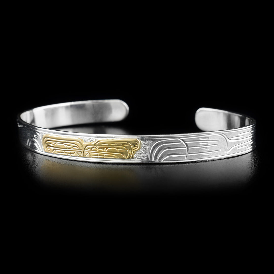 This gold and silver bracelet depicts the head of an orca and a bear facing one another in the center, made from gold. To the right is the bear's paw.