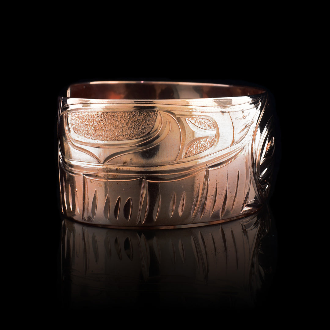 This copper cuff bracelet has a single, wide band with a gap in the back. The baby Hummingbird is depicted in the center with its head surrounded by an oval designs representing the womb. Where its beak meets the womb on the right, there are petals designs. The head of the mother Hummingbird is carved on the right side and the left depicts her outstretched wing.