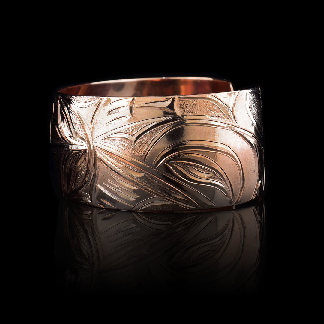 This copper cuff bracelet has a single, wide band with a gap in the back. The baby Hummingbird is depicted in the center with its head surrounded by an oval designs representing the womb. Where its beak meets the womb on the right, there are petals designs. The head of the mother Hummingbird is carved on the right side and the left depicts her outstretched wing.