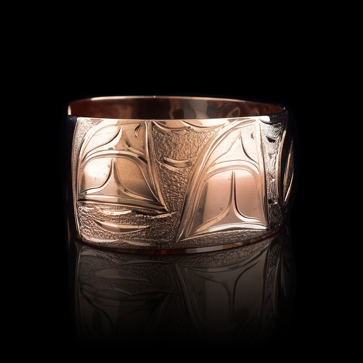 This copper cuff bracelet has a single, side band with a gap in the back. There is a baby Orca depicted in the womb with its mother watching over is on one side of the piece and a two dorsal fins in the ocean depicted on the other side. This piece represents family and community.