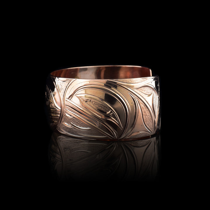 This copper cuff bracelet has a single, side band with a gap in the back. There is a baby Orca depicted in the womb with its mother watching over is on one side of the piece and a two dorsal fins in the ocean depicted on the other side. This piece represents family and community.