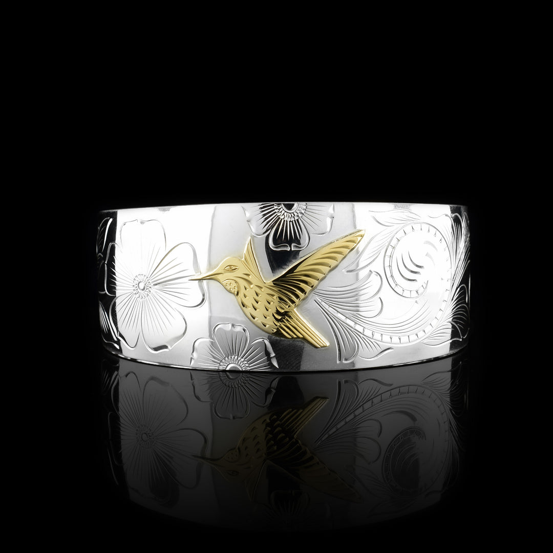 This sterling silver and gold hummingbird bracelet cuff has a single, wide band with a gap in the back. The band tapers slightly in the back. There is a gold hummingbird in the center with carvings detailing its beak, eye and feather patterns. Both wings are outstretched in flight. There are flowers and floral designs carved into the silver around the bird.