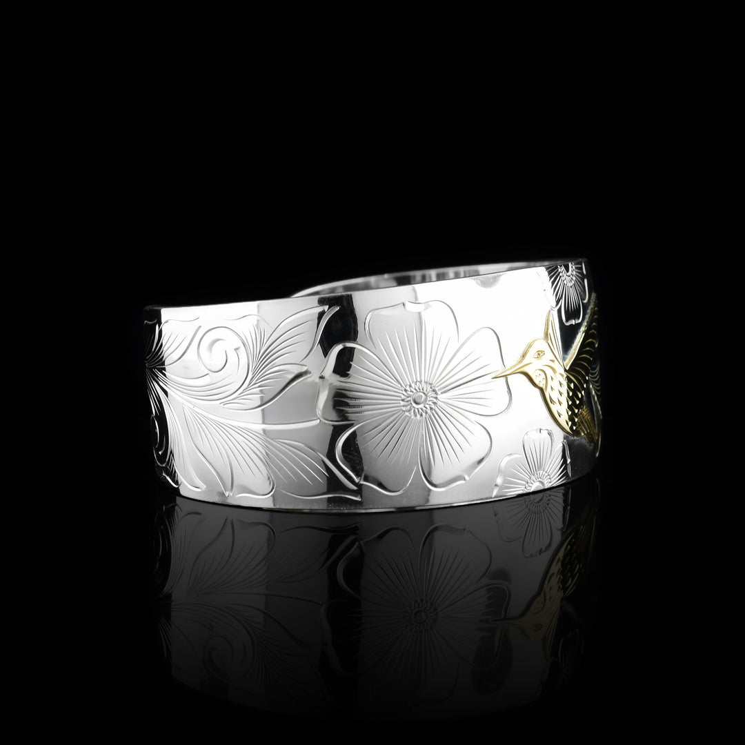 This sterling silver and gold hummingbird bracelet cuff has a single, wide band with a gap in the back. The band tapers slightly in the back. There is a gold hummingbird in the center with carvings detailing its beak, eye and feather patterns. Both wings are outstretched in flight. There are flowers and floral designs carved into the silver around the bird.