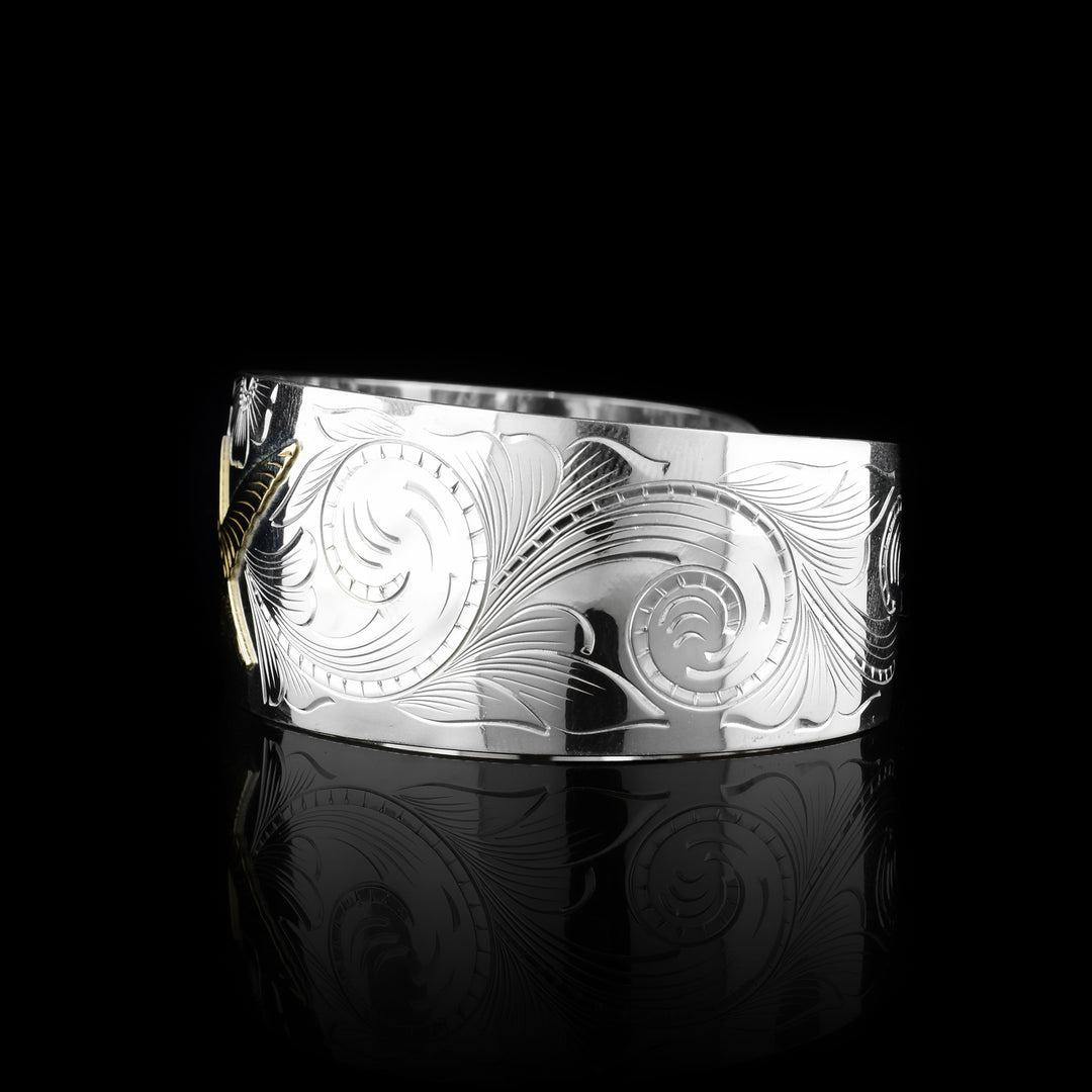 This sterling silver and gold hummingbird bracelet cuff has a single, wide band with a gap in the back. The band tapers slightly in the back. There is a gold hummingbird in the center with carvings detailing its beak, eye and feather patterns. Both wings are outstretched in flight. There are flowers and floral designs carved into the silver around the bird.