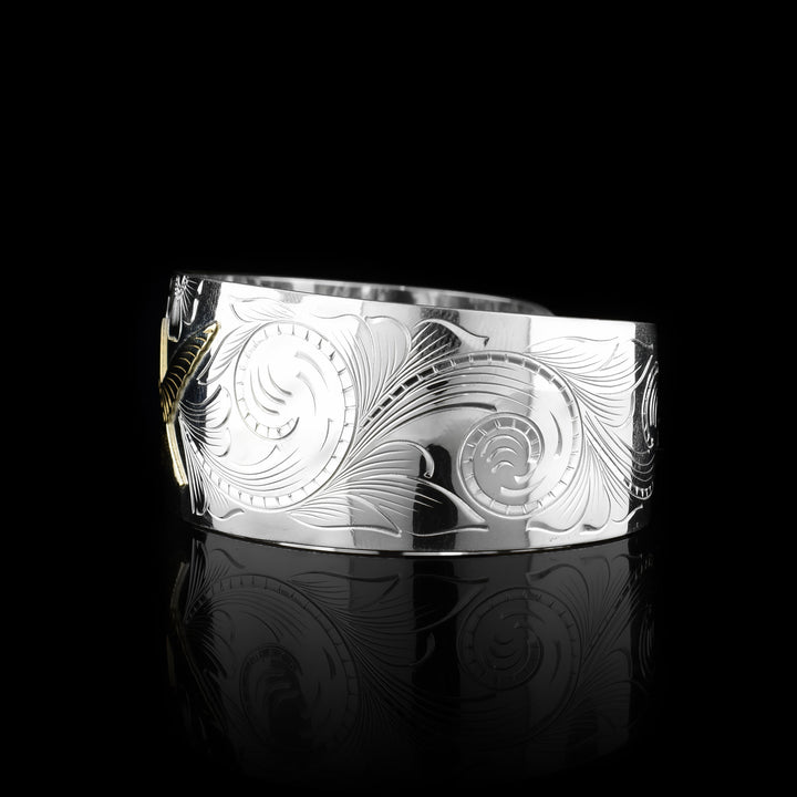 This sterling silver and gold hummingbird bracelet cuff has a single, wide band with a gap in the back. The band tapers slightly in the back. There is a gold hummingbird in the center with carvings detailing its beak, eye and feather patterns. Both wings are outstretched in flight. There are flowers and floral designs carved into the silver around the bird.