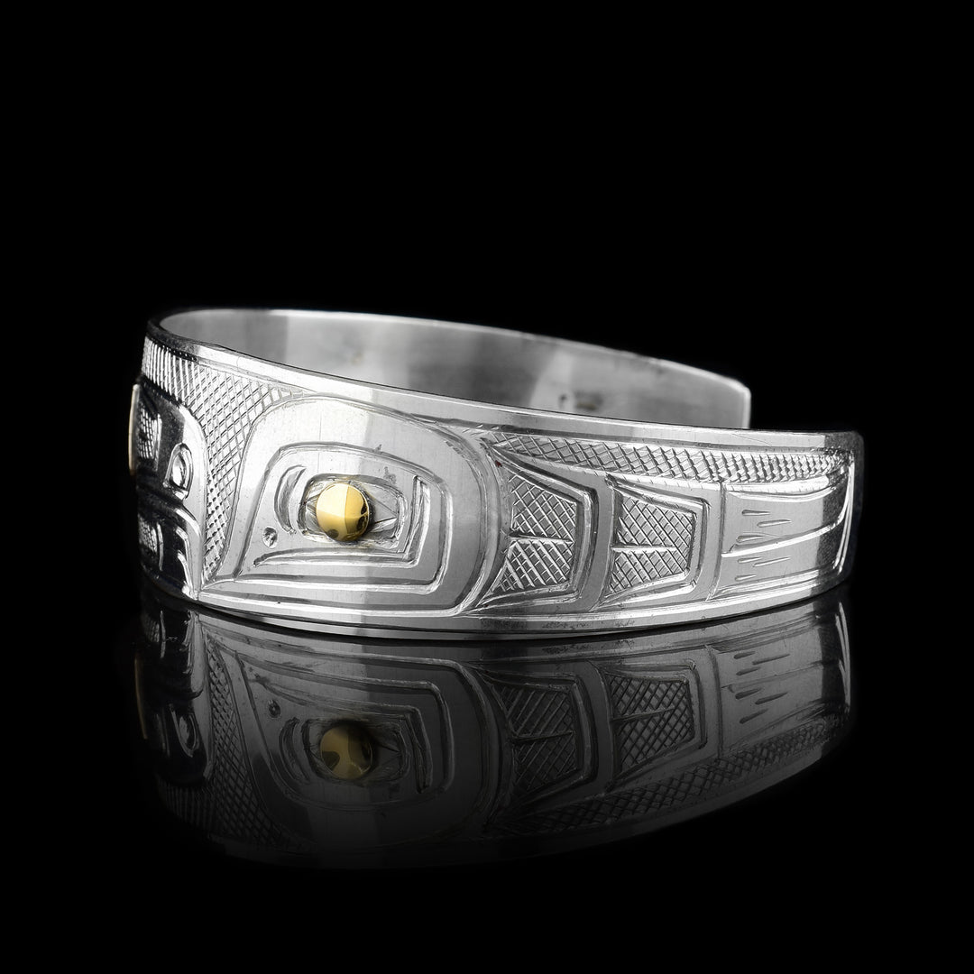 This Salmon and Eagle bracelet cuff is made from sterling silver, 14k gold and has a single, tapered band with a gap in the back. There is a depiction of the Eagle and the Salmon carved into the band. The eyes of the legends are made with gold studs.