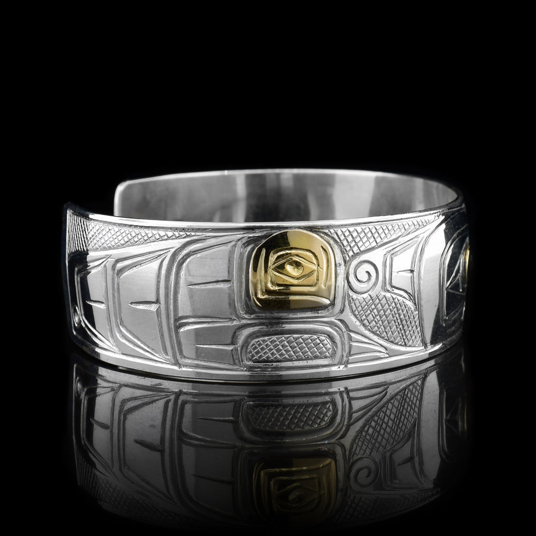 This Thunderbird bracelet is made from sterling silver and 14K gold with a wide band that has a gap in the back. There is a depiction of the Thunderbird carved into the surface of the band and there are gold embellishments on the wings and the eye of it.