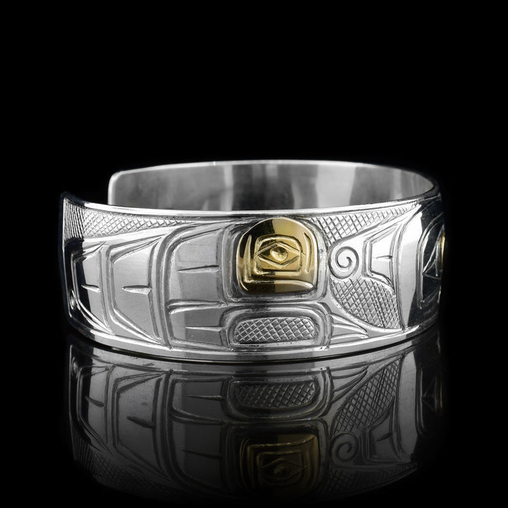 This Thunderbird bracelet is made from sterling silver and 14K gold with a wide band that has a gap in the back. There is a depiction of the Thunderbird carved into the surface of the band and there are gold embellishments on the wings and the eye of it.