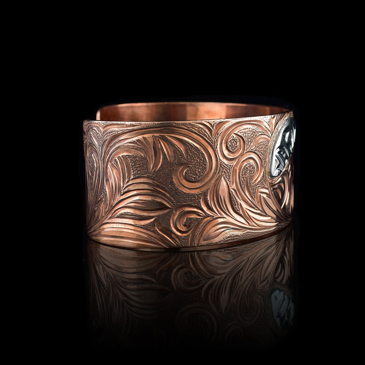 This Bear bracelet has a wide band made from copper and with a gap in the back. In the center, the baby Bear is depicted with its head facing left and its paws curlced up in front of it. Around the baby there is an oval designs which represents the womb and there is a sterling silver oval with carvings on it to the opposite side of the baby. On the right end of the band, the mother bear is depicted with a side profile of her head which is tilted upwards. On the other side, there are floral designs