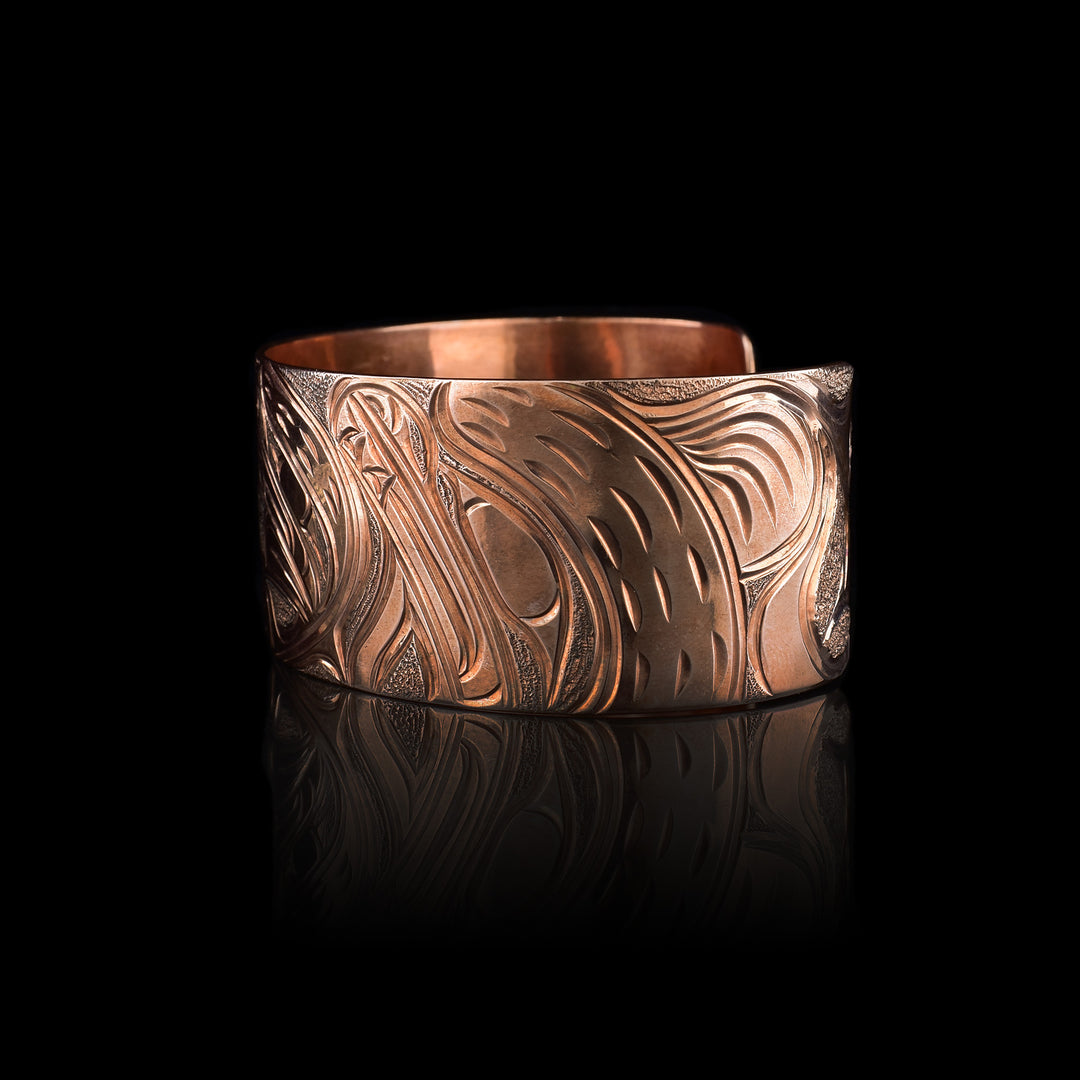 This Bear bracelet has a wide band made from copper and with a gap in the back. In the center, the baby Bear is depicted with its head facing left and its paws curlced up in front of it. Around the baby there is an oval designs which represents the womb and there is a sterling silver oval with carvings on it to the opposite side of the baby. On the right end of the band, the mother bear is depicted with a side profile of her head which is tilted upwards. On the other side, there are floral designs