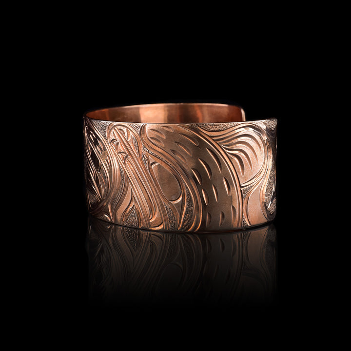 This Bear bracelet has a wide band made from copper and with a gap in the back. In the center, the baby Bear is depicted with its head facing left and its paws curlced up in front of it. Around the baby there is an oval designs which represents the womb and there is a sterling silver oval with carvings on it to the opposite side of the baby. On the right end of the band, the mother bear is depicted with a side profile of her head which is tilted upwards. On the other side, there are floral designs