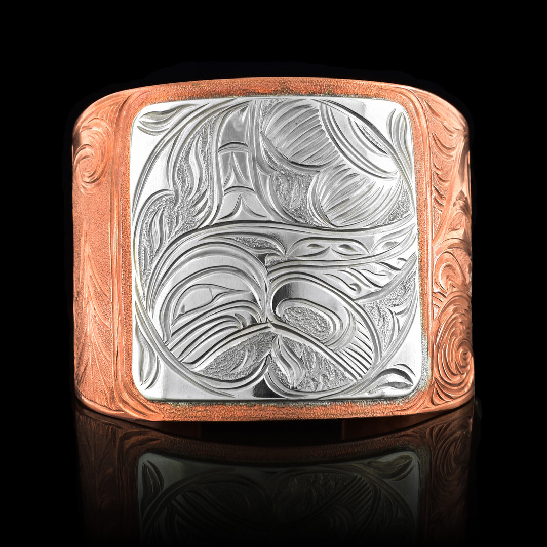 This large copper cuff bracelet has a very wide copper band with a gap in the back. The band is made from copper and tapers to be thinner at the end. There is a sterling silver square in the center of the band with a full body Orca done in a side profile carved into it. On one end of the band there are dorsal fins emerging from waves and on the other the wind spirt can be seen above forest trees.