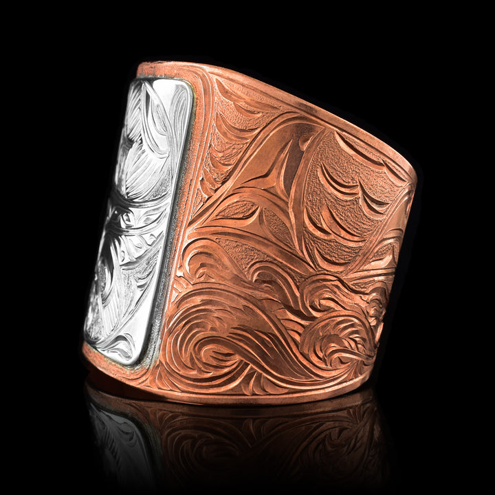 This large copper cuff bracelet has a very wide copper band with a gap in the back. The band is made from copper and tapers to be thinner at the end. There is a sterling silver square in the center of the band with a full body Orca done in a side profile carved into it. On one end of the band there are dorsal fins emerging from waves and on the other the wind spirt can be seen above forest trees.