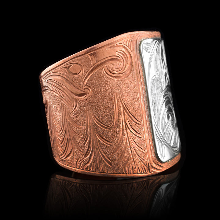 This large copper cuff bracelet has a very wide copper band with a gap in the back. The band is made from copper and tapers to be thinner at the end. There is a sterling silver square in the center of the band with a full body Orca done in a side profile carved into it. On one end of the band there are dorsal fins emerging from waves and on the other the wind spirt can be seen above forest trees.