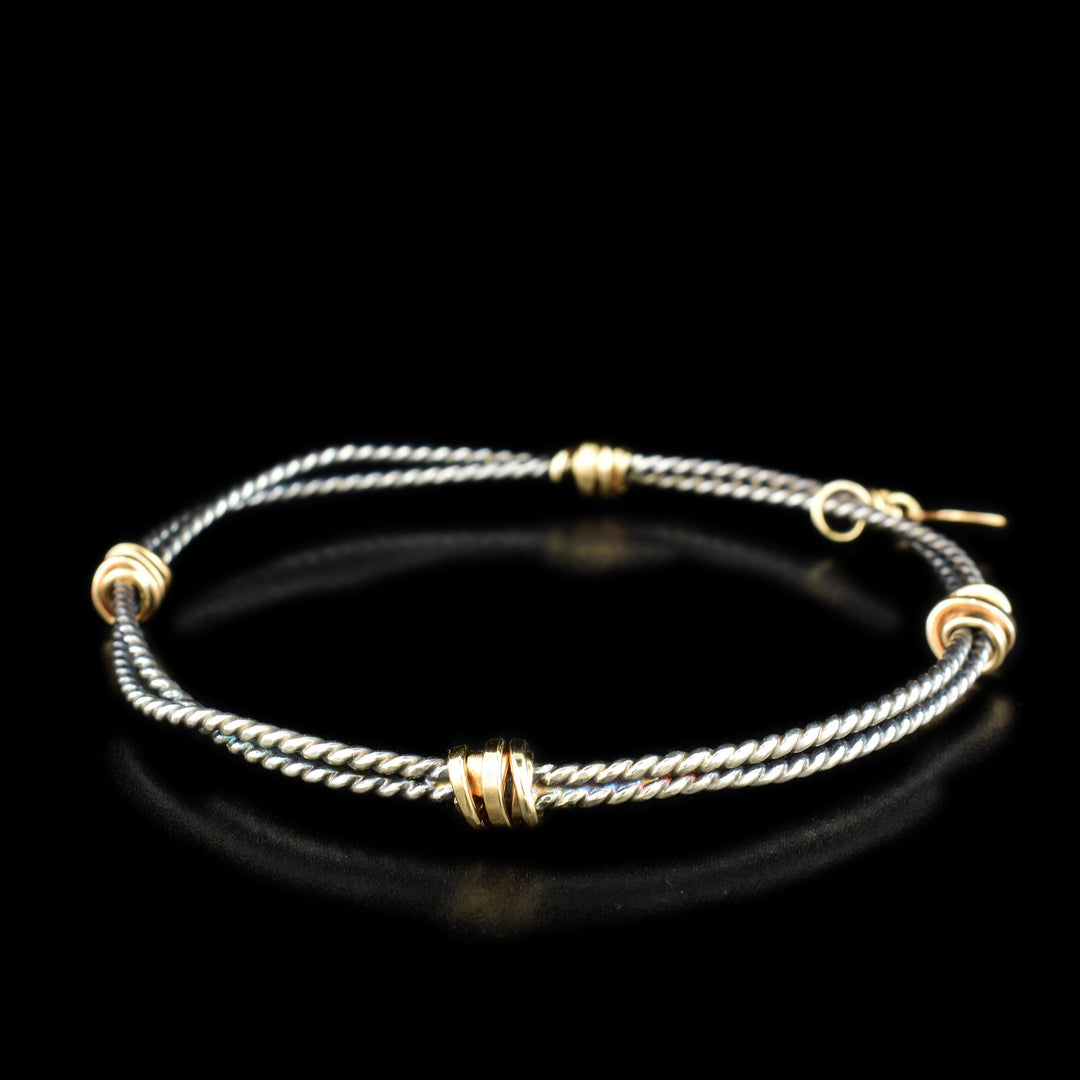 This twisted bangle bracelet features two twisted cords of silver that have been joined together with four gold-filled accents placed evenly throughout. This bracelet features a small gold-filled charm.