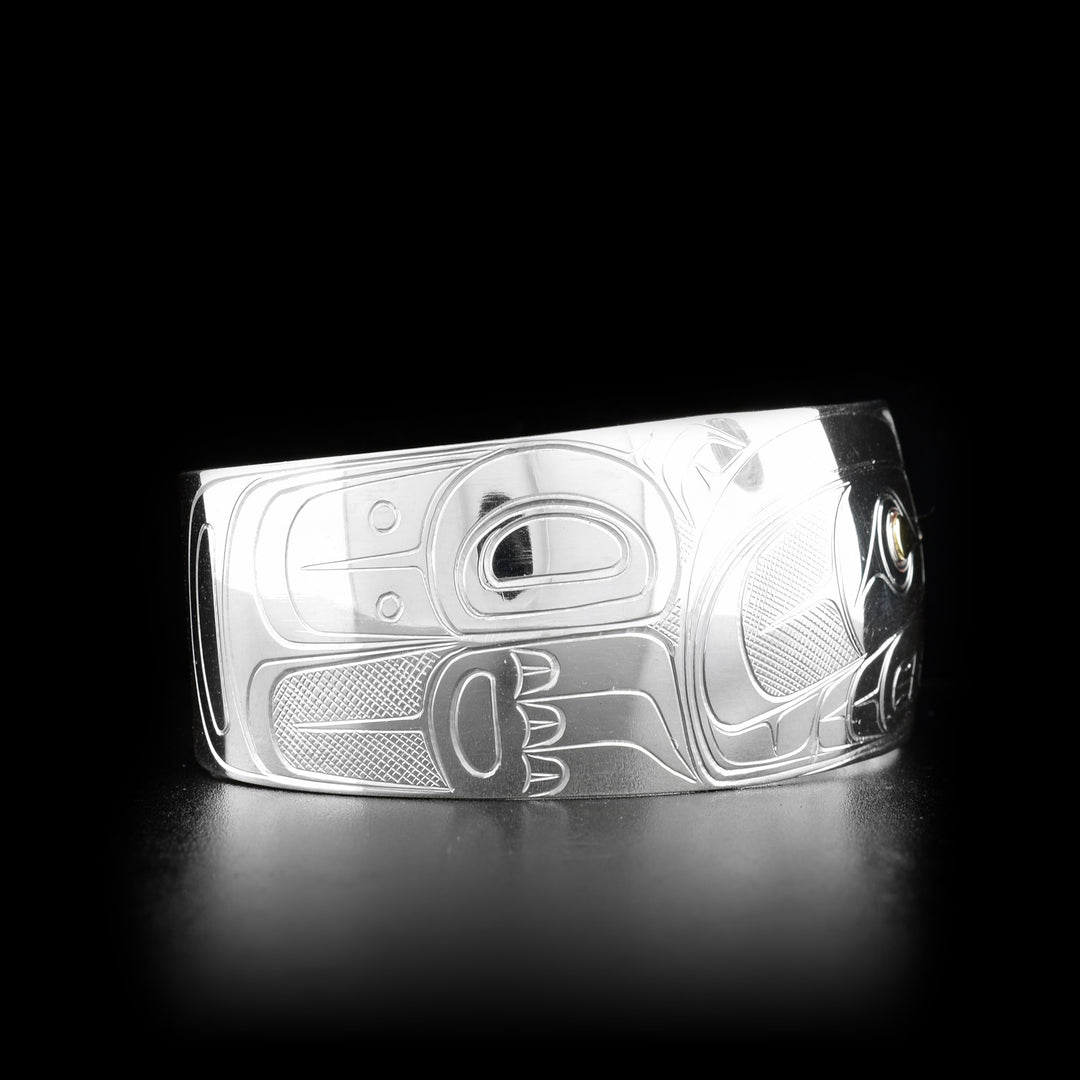 This silver and gold bracelet is made from sterling silver and 14K gold and has a wide band that tapers slightly. There is a depiction of the Bear carved into the surface of the band and the eye of the Bear is made from 14K gold.