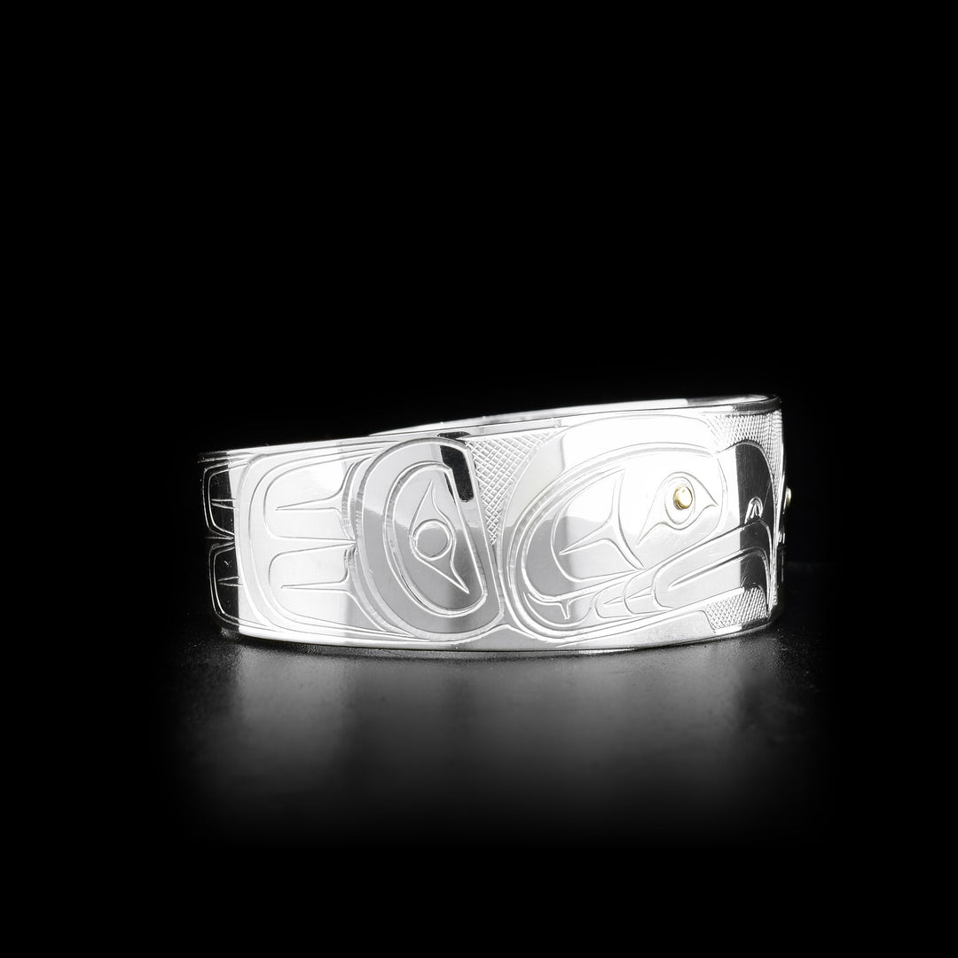 This gold and silver bracelet cuff is made from sterling silver and 14K gold and has a wide band with a gap in the back. There are depictions of the Eagle and the Orca carved into the surface of the band and the eyes of the Legends are made from 14K gold.